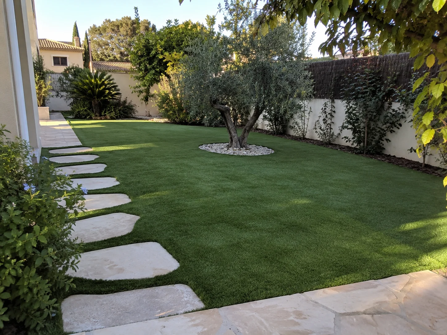 Jardín con césped artificial, olivo y camino de pasos